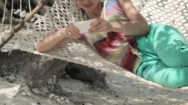 Młoda kobieta tekstylny, wysyłanie SMS-ów na smartphone, leżącego na hamaku — Wideo stockowe