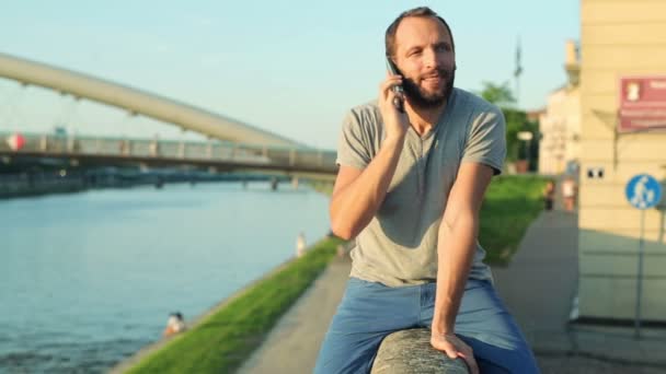 Разговаривая по сотовому, человек сидит на стене у реки — стоковое видео