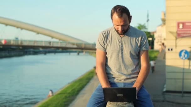 Man med laptop sitter på väggen vid floden — Stockvideo