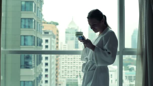 Женщина со смартфоном у окна — стоковое видео