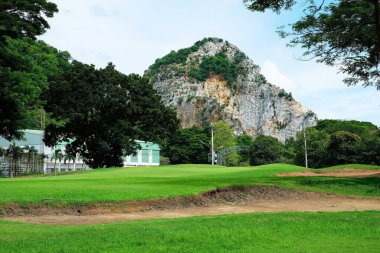 Yeşil çimen, kum sığınağı, ağaçlar ve mavi gökyüzünün altındaki kayalık dağ manzarası.