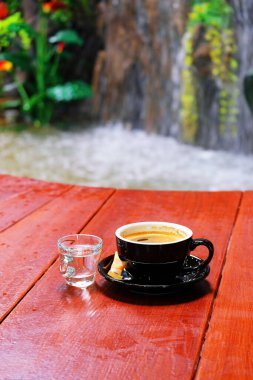 Kırmızı ahşap zeminde bir bardak suyla siyah bir fincanda sıcak kahve. Bulanık adam şelale arkaplanı yapmadan önce..