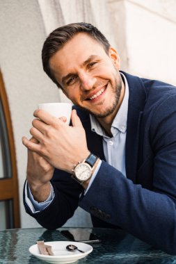 Adam kahveyi seviyor. Kafede bir fincan kahveyle yakışıklı bir adam. Erkeğin sabah yaşam tarzı. Adam yazlık terasta oturmuş kahve içiyor ve gülümsüyor.