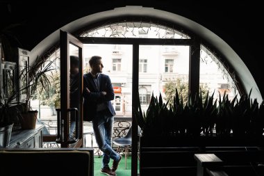 Yakışıklı erkek model kafede poz veriyor. Sakallı adam takım elbiseli ve pencerenin yanında gülümsüyor.