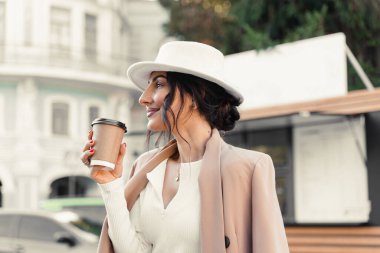 İş kadını kahve dükkanının yanında kahve içiyor. Beyaz şapka, elbise ve ceket giymiş bir kız.
