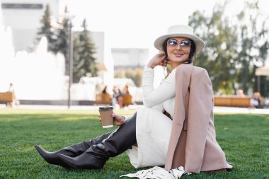 Çimlerin üzerinde bir fincan kahve oturağı olan manken. Çekici kadın kırbaç, şapka, elbise, ceket ve güneş gözlüğü takıyordu.