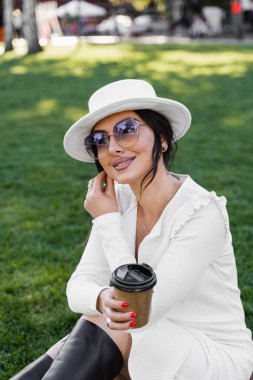 Çimlerin üzerinde bir fincan kahvesi olan çekici bir kadın. Beyaz giyimli resmi model kahvenin tadını çıkar.