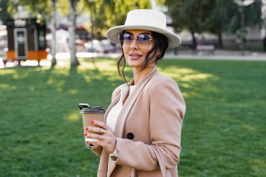 Akıllı telefon ve kahve fincanı olan blog yazarının sokak tarzı portresi. Beyaz şapka, elbise ve ceket giymiş bir kız.