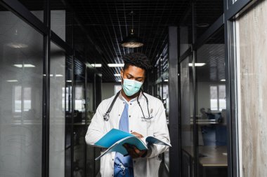 Siyahi tıp öğrencisi kitap okuyor ve sınava hazırlanıyor. Tıbbi giysili ve maskeli Afrikalı notlara bakıyor.