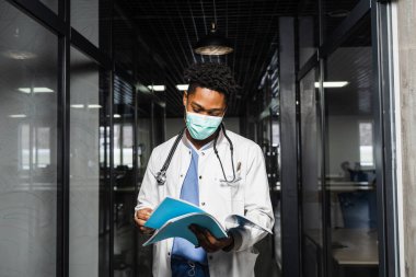 Siyahi tıp öğrencisi kitap okuyor ve sınava hazırlanıyor. Tıbbi giysili ve maskeli Afrikalı notlara bakıyor.