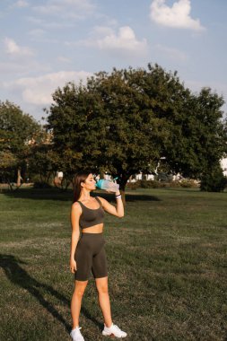 Atletik formda bir Asyalı kız yeşil parktaki bir şişeden su içiyor. Eğitimden sonra dinlen.
