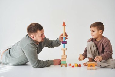 Önemseyen baba, beyaz arka planda oğlunun yerde oynamasına yardım eder. Baba ve çocuk renkli ahşap tuğlalardan bir kule inşa eder ve birlikte eğlenirler.