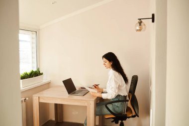 Çekici kız öğrenci evde dizüstü bilgisayar kullanarak online çalışıyor. Çevrimiçi eğitim ve uzaktan öğrenme. Kıvırcık saçlı kız internette beyaz gömlek giyiyor.