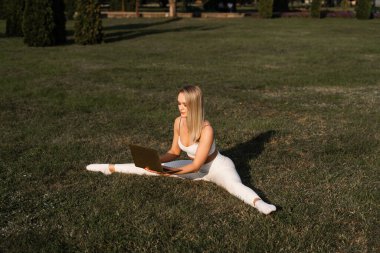 Zinde bir sarışın çimenlerde bacağını ikiye ayırıyor. Parkta dizüstü bilgisayarla antrenman yapıyordum. Spor yaşam tarzı