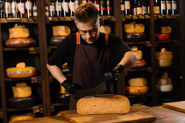 チーズショップのハンサムな男労働者はナイフで限られたグダを切りました チーズショップで働くチーズソムリエ チーズ製品の広告のための創造的なアイデア — ストック写真