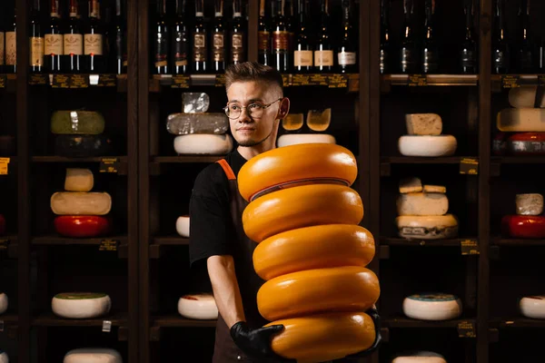 Big yellow wheels of cheese in hands of handsome sommelier. Worker of food cheese store carrying rounds of cheese