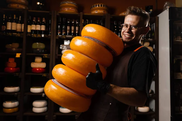 Big yellow wheels of cheese in hands of handsome sommelier. Worker of food cheese store carrying rounds of cheese