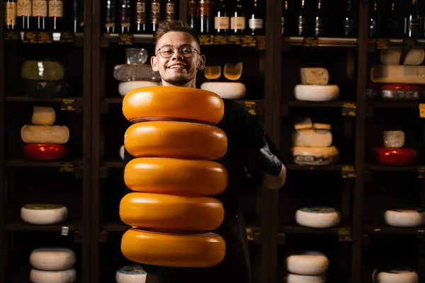 Handsome cheese sommelier with yellow wheels of cheese in shop. Worker of food cheese store carrying rounds of cheese