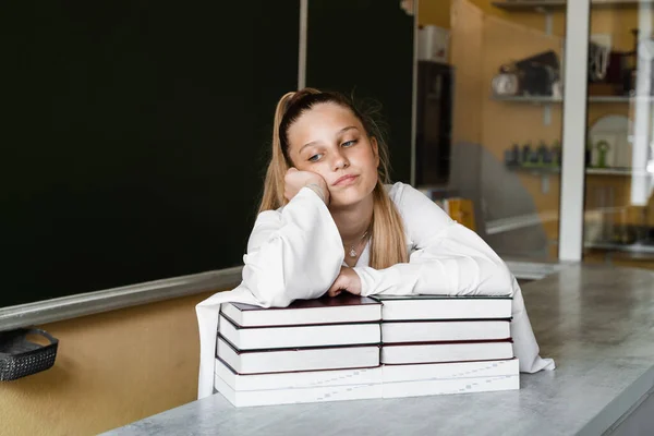 Tired and frustrated schoolgirl lies on books at blackboard in school. School education. The girl is tired of teaching homework