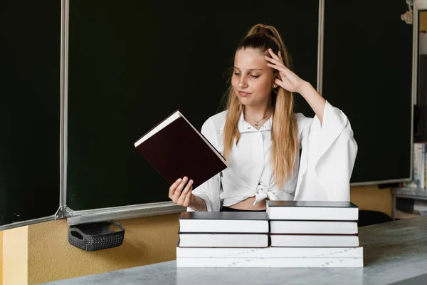 School education. Schoolgirl looks at the books and gets upset because of the large amount of homework. Sad child posing with book at blackboard in school