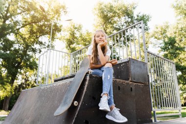 Kaykay tahtası ile rampada oturan kız çocuğu. Çocuklar için spor malzemeleri. Kaykay parkında kaykay yapan aktif bir genç.