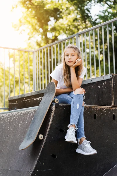 Child girl sitting with skate board on sport ramp. Sports equipment for kids. Active teenager with skate board on skate park playground