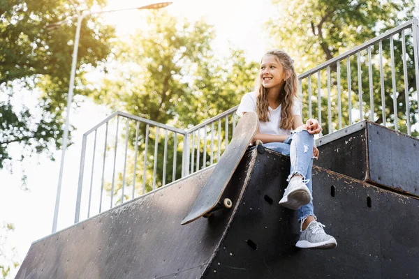 Child girl sitting with skate board on sport ramp. Sports equipment for kids. Active teenager with skate board on skate park playground