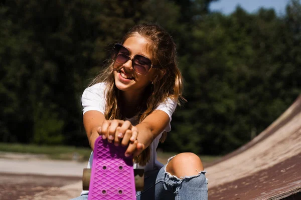 Happy smiling girl with skate board sitting on skate playground and having fun. Extreme sport lifestyle. Laughing child with skate board posing on sport ramp