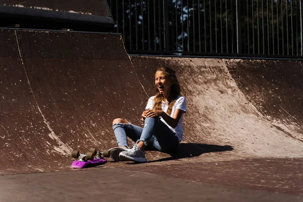 Happy smiling girl with skate board sitting on skate playground and having fun. Extreme sport lifestyle. Laughing child with skate board posing on sport ramp