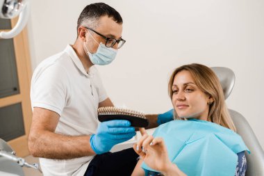 Diş hekimi, diş sağlığı kliniğindeki kadın hastalara diş beyazlatma için diş tonu rehberliği gösteriyor. Diş hekimliği. Kadın kaplamalara bakıyor ya da diş implantı yerleştiriyor. Diş renkleri doktorun ellerinde eşleşiyor.