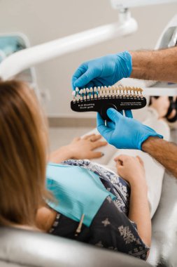 Kaplamalara bakan ya da diş implantı takan bir kadın. Doktorun ellerine eşleşen diş renkleri. Diş hekimliği. Diş hekimi, diş sağlığı kliniğinde kadın hastalara diş beyazlatma için diş tonları rehberliği gösteriyor