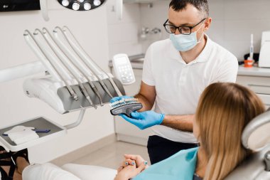 Diş hekimi, diş sağlığı kliniğindeki kadın hastalara diş beyazlatma için diş tonu rehberliği gösteriyor. Diş hekimliği. Kadın kaplamalara bakıyor ya da diş implantı yerleştiriyor. Diş renkleri doktorun ellerinde eşleşiyor.