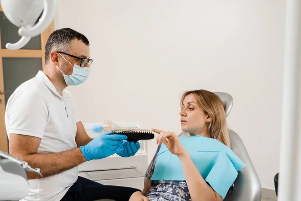 Consultation with dentist about teeth whitening and showing girl patient teeth color shades guide in dental clinic. Dentistry. Woman looking at teeth color matching samples in doctor hands