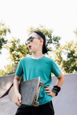 Fashion child boy in glasses posing with skate board on sport playground. Active teenager with sport equipment on sport ramp. Extreme lifestyle