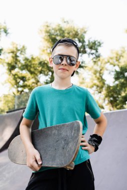 Fashion child boy in glasses posing with skate board on sport playground. Active teenager with sport equipment on sport ramp. Extreme lifestyle