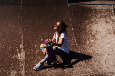Çocuk model gözlüklü, paten sahasında oturmuş ve paten kayarken poz veriyor. Spor rampasında ekstrem yaşam tarzından hoşlanan aktif kız çocuğu