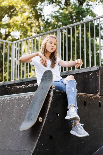 Having fun with skate board. Funny child girl with skate sitting on sport ramp, smiling and grimacing on skate playground. Active teenager posing with skate board. Extreme lifestyle