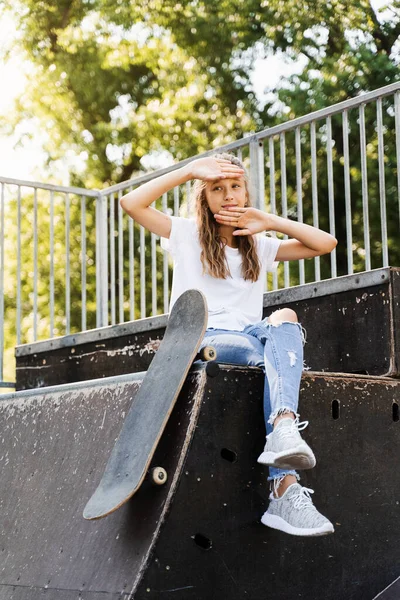 Having fun with skate board. Funny child girl with skate sitting on sport ramp, smiling and grimacing on skate playground. Active teenager posing with skate board. Extreme lifestyle