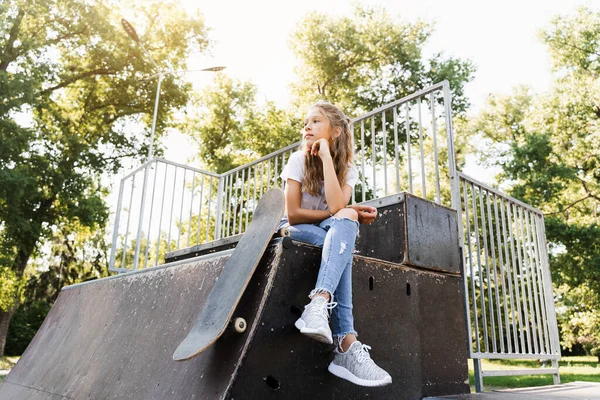 Child girl sitting with skate board on sport ramp. Sports equipment for kids. Active teenager with skate board on skate park playground