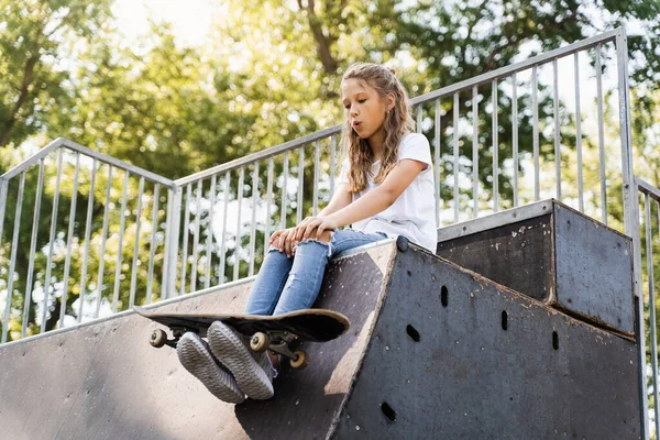 Happy smiling girl with skate board sitting on skate playground and having fun. Extreme sport lifestyle. Laughing child with skate board posing on sport ramp