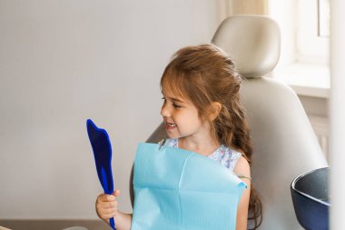 Çocuk dişçide aynaya bakıyor. Mutlu çocuk diş hekimi. Diş tedavisi. Çekici kız dişçi ofisinde oturuyor ve gülümsüyor.