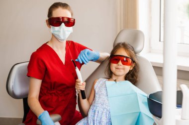 Çocuk diş hekimi ve kırmızı gözlüklü çocuk hasta. Fotopolimer diş dolgusunun UV aydınlatmasına hazırlık. Diş hekimi çocuk hastaları tedavi eder ve kaldırır.