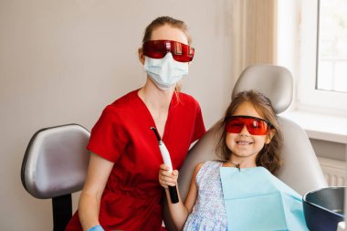 Çocuk diş hekimi ve kırmızı gözlüklü çocuk hasta. Fotopolimer diş dolgusunun UV aydınlatmasına hazırlık. Diş hekimi çocuk hastaları tedavi eder ve kaldırır.