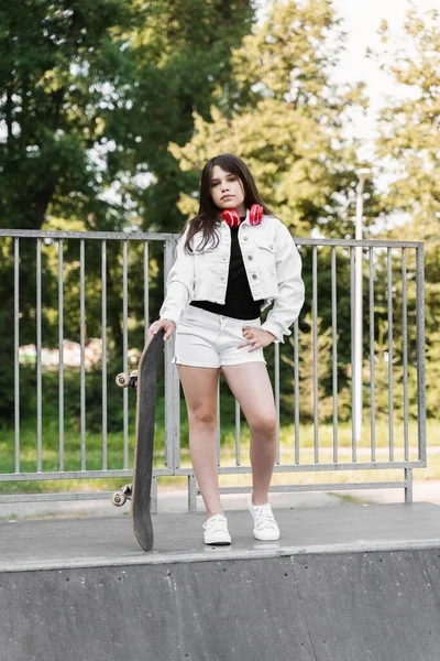 Child girl with skate board and headphones posing on skate sport ramp. Sports equipment for kids. Active teenager with skate board on skate park playground