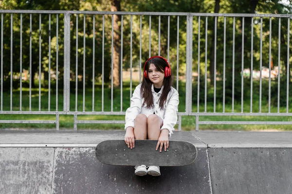 Active child girl with skate board and listen to music with headphones and sitting on skate playground and smile. Extreme sport lifestyle. Smiling child with skateboard posing on sport ramp