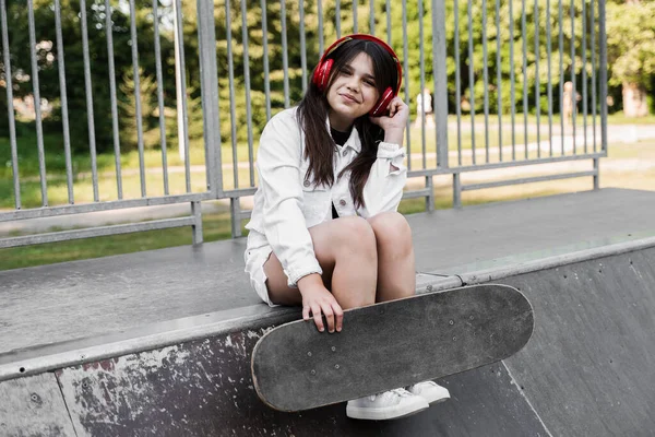 Active child girl with skate board and listen to music with headphones and sitting on skate playground and smile. Extreme sport lifestyle. Smiling child with skateboard posing on sport ramp