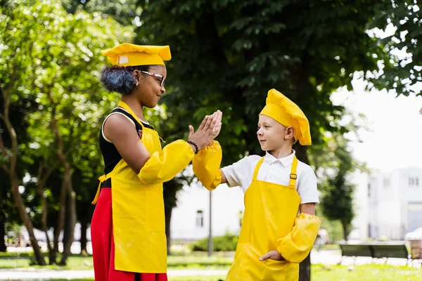 Clap hands and give five. Black african and caucasian cook child in yellow chefs hat and apron clap each others hands and give five. Multiracial friends smiling and having fun together