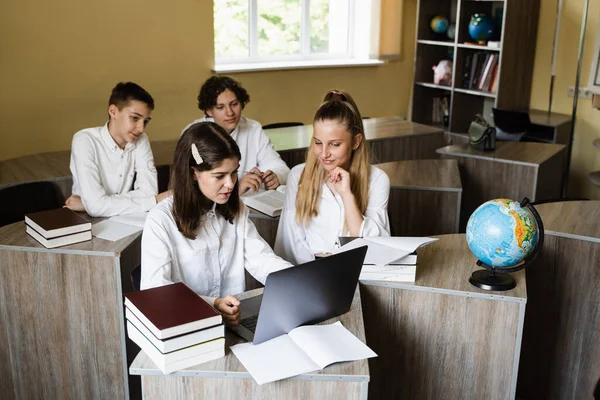 Children in school at geography lesson with globe study online on laptop and discuss different countries. Education concept. Pupils at school communicating with classmates online