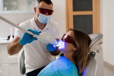 Artificial teeth whitening. Woman in red protective glasses patient do UV teeth whitening procedure. Dental ultraviolet whitening treatment with light, fluoride and laser