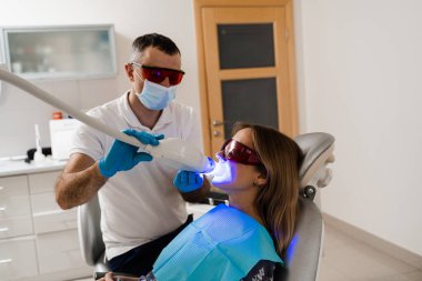 Artificial teeth whitening. Woman in red protective glasses patient do UV teeth whitening procedure. Dental ultraviolet whitening treatment with light, fluoride and laser